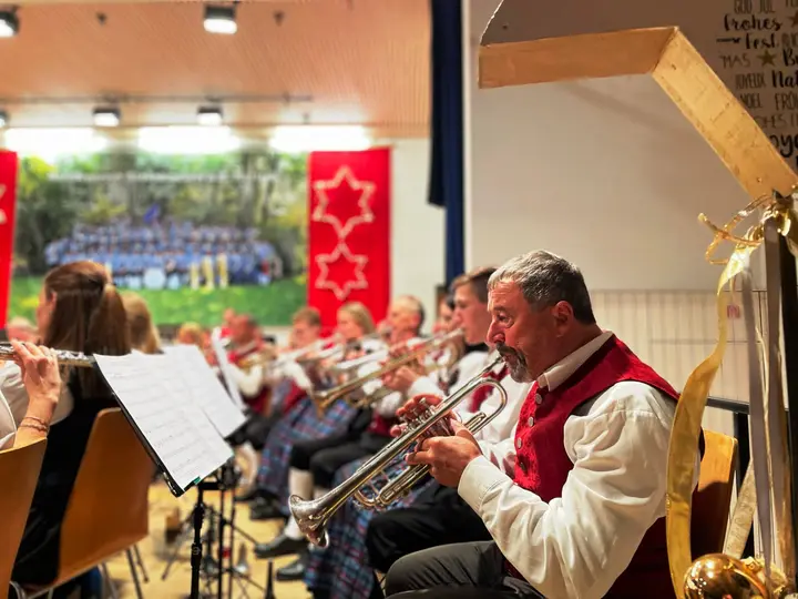 Musikerinnen und Musiker in Tracht spielen Trompete und Querflöte in der weihnachtlich geschmückten Halle, im Hintergrund ein großes Gruppenfoto des Vereins und rote Banner mit Sternen