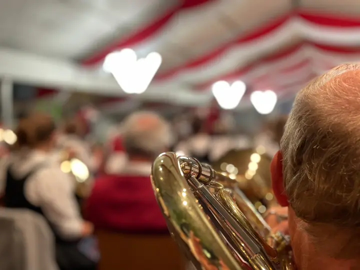 Blick über die Schulter eines Musikers auf sein glänzendes Blechblasinstrument, dahinter unscharf die Kapelle im rot-weiß geschmückten Festzelt mit herzförmigen Lichtern