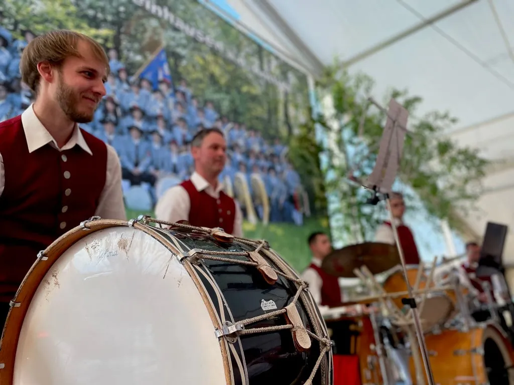 Ein Musiker in roter Weste lächelt hinter der großen Trommel, im Hintergrund das Schlagzeug, weitere Musiker und ein Banner mit einem Gruppenfoto des Vereins
