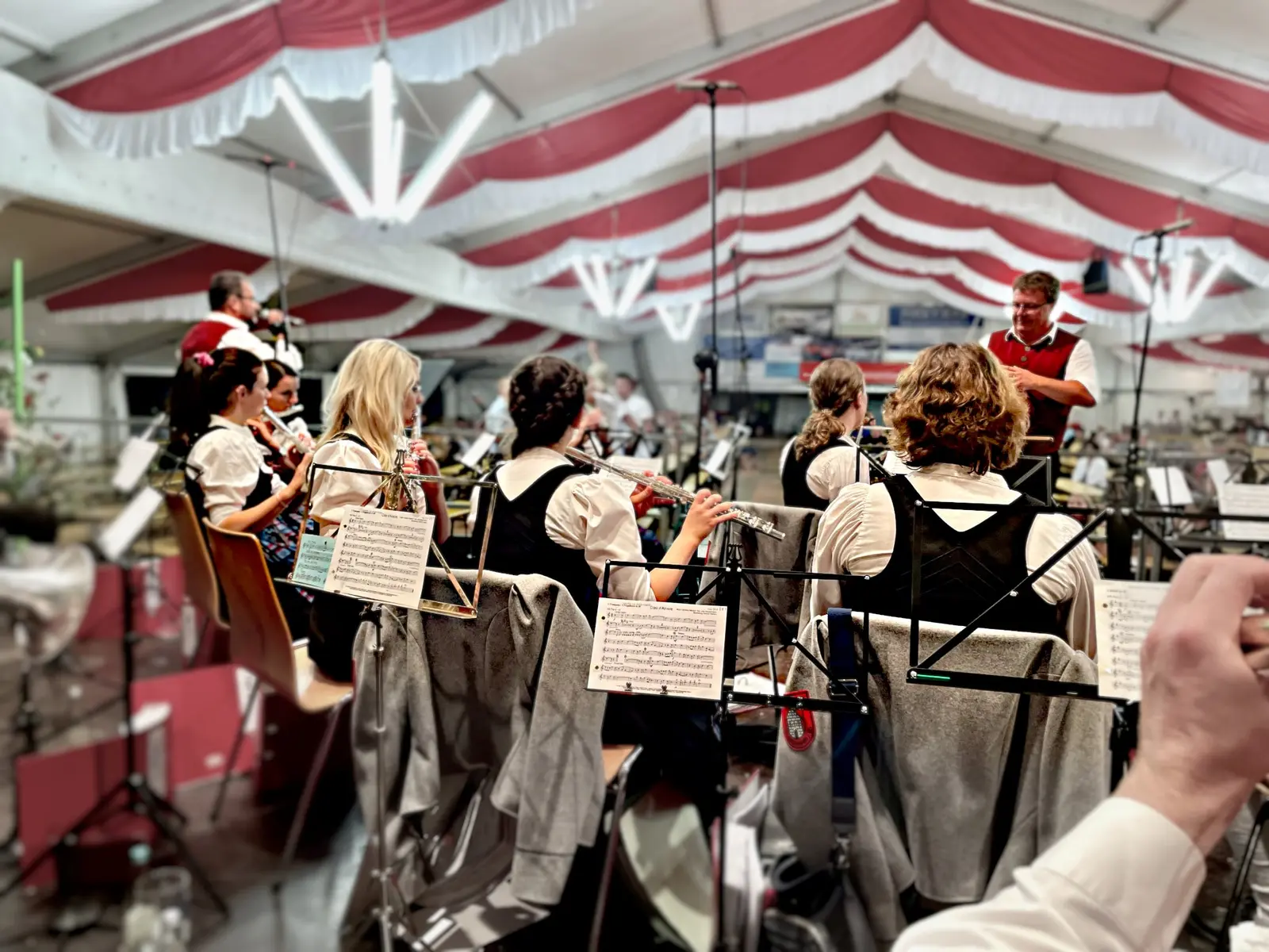 Blick über die Schultern der Musikkapelle im rot-weiß geschmückten Festzelt: Musikerinnen in Tracht spielen Querflöte, rechts steht der Dirigent in roter Weste