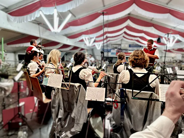 Blick über die Schultern der Musikkapelle im rot-weiß geschmückten Festzelt: Musikerinnen in Tracht spielen Querflöte, rechts steht der Dirigent in roter Weste