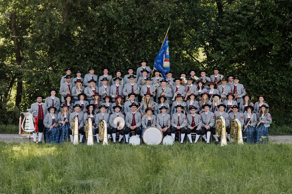 Gruppenbild der Kapelle in Tracht mit Instrumenten und Vereinsfahne auf einer Wiese vor Bäumen