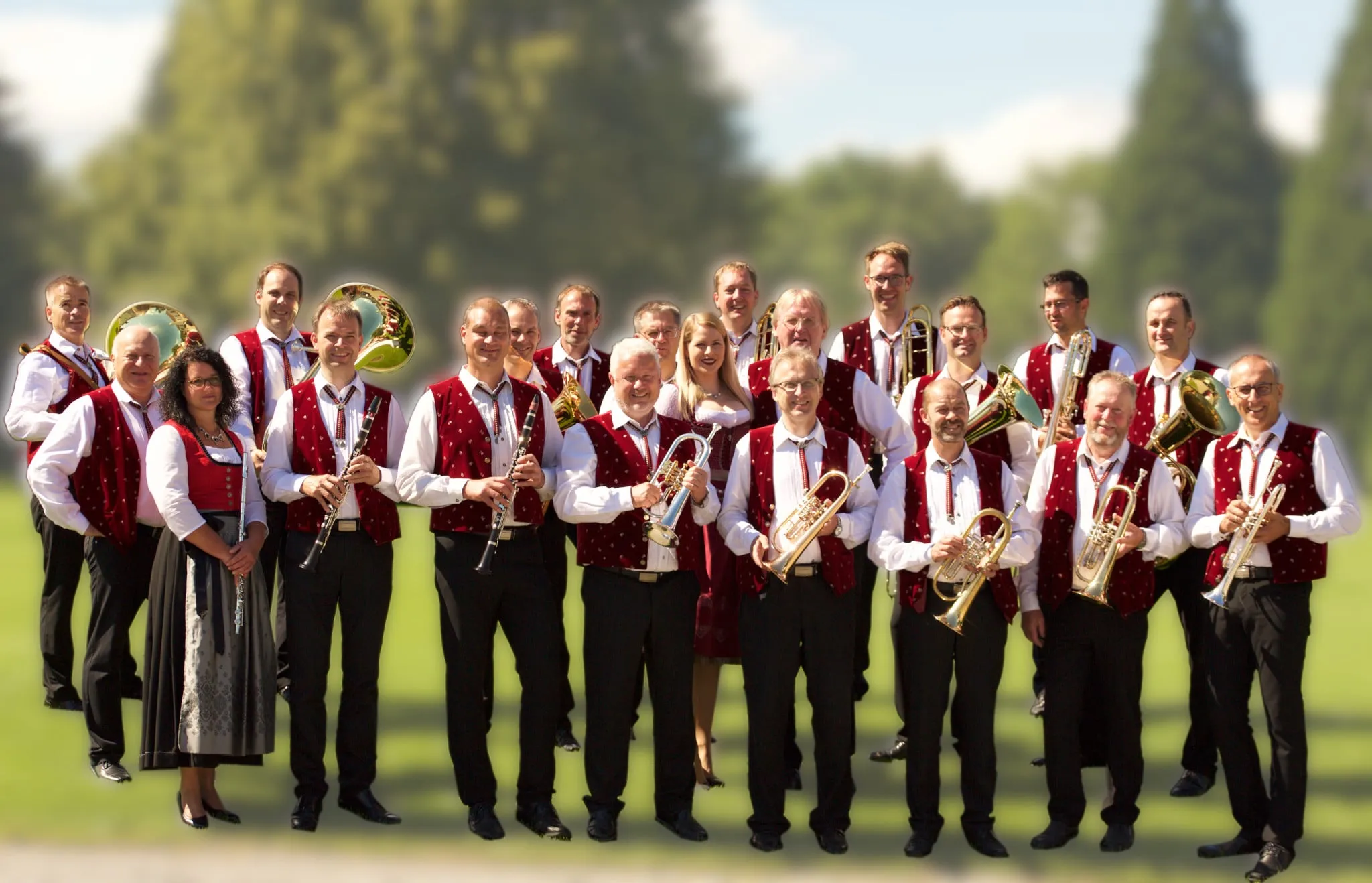 Gruppenfoto der Oberschwäbischen Dorfmusikanten auf einer sonnigen Wiese: Musiker in roten Westen und weißen Hemden mit ihren Blasinstrumenten, dazu eine Flötistin und eine Sängerin im Dirndl