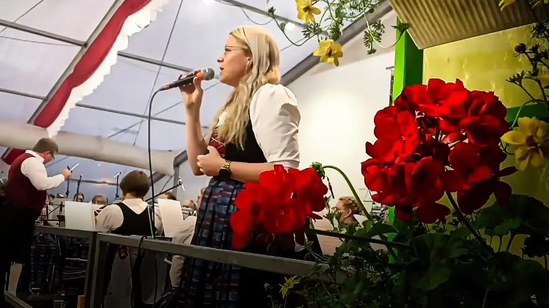 Eine Sängerin in Tracht singt im Festzelt ins Mikrofon, im Vordergrund rote Geranien, im Hintergrund die Blaskapelle mit ihrem Dirigenten