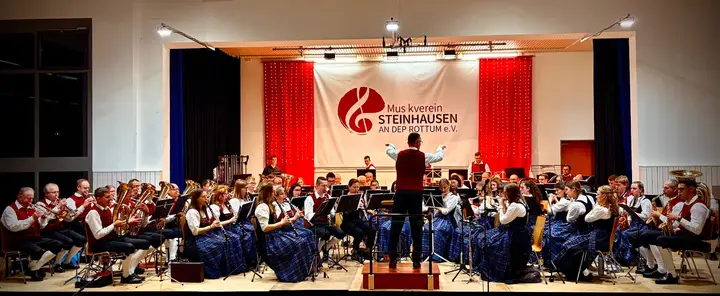 Unser Blasorchester in Tracht auf der Bühne der Mehrzweckhalle, in der Mitte der Dirigent auf dem Podest, dahinter das Vereinsbanner zwischen roten Vorhängen mit Lichterketten
